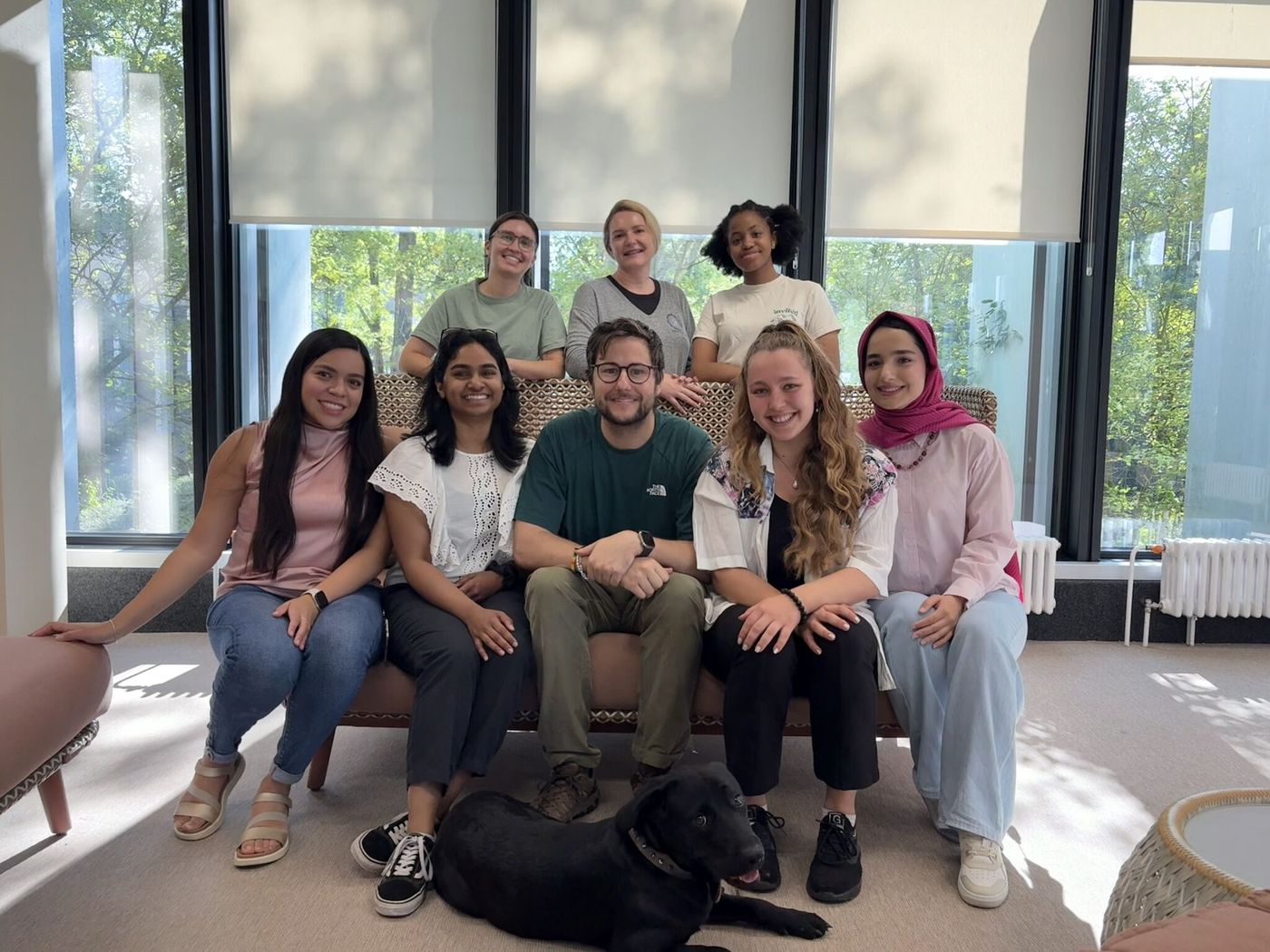 The Schulz Squad group photo: eight people on and behind a sofa by tall windows, with a black dog lying in front